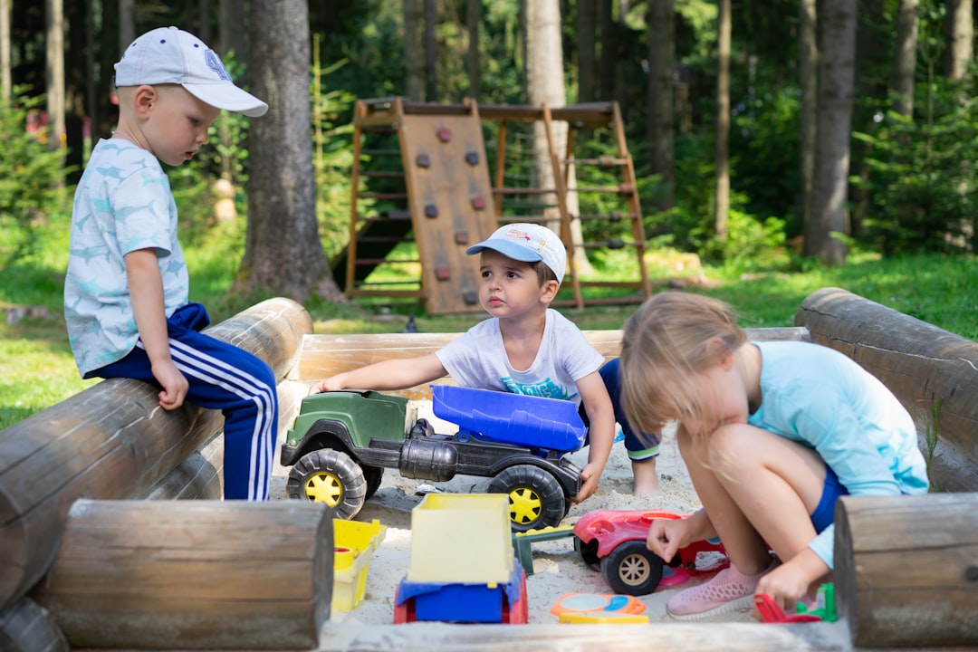 Three young children in caps playing with toy trucks in a wooden sandbox at an outdoor daycare playground surrounded by trees