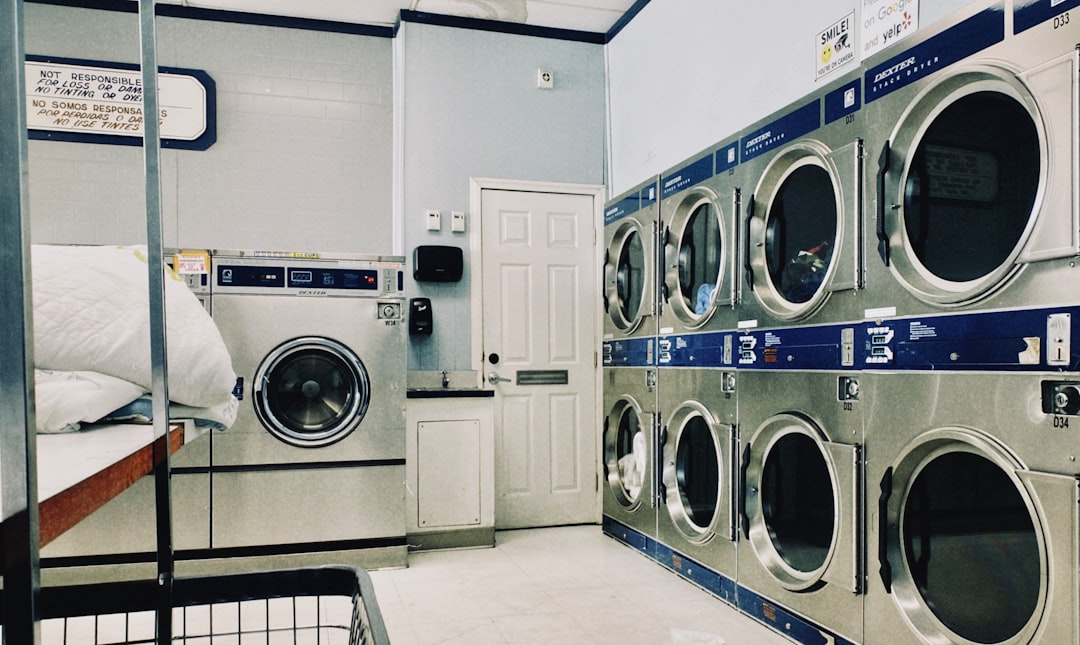 Interior of a laundromat with a wall of blue Dexter stack dryers on the right, a large front-load washer, a white door, and a warning sign on the left wall