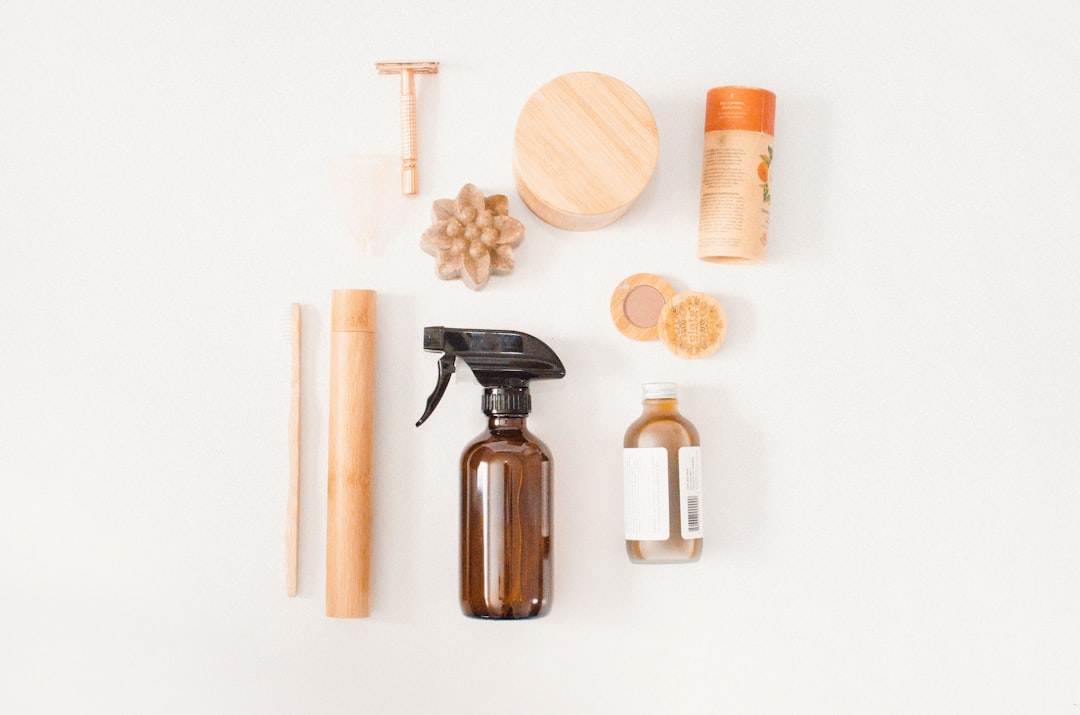 Flat lay of natural cleaning and grooming supplies on a white background — amber glass spray bottle, wooden brush, bamboo containers, a soap flower, and a safety razor