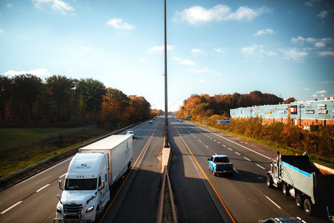 White Freightliner semi hauling a dry van on a multi-lane highway past an outlet mall on an autumn afternoon